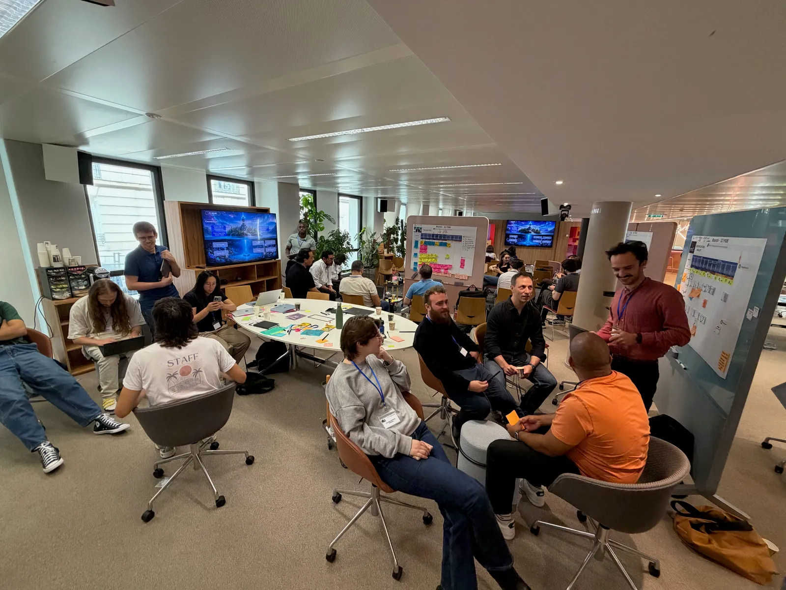Plusieurs groupes réunis dans un espace de travail, autour de tables, d’ordinateurs et de tableaux de notes.
